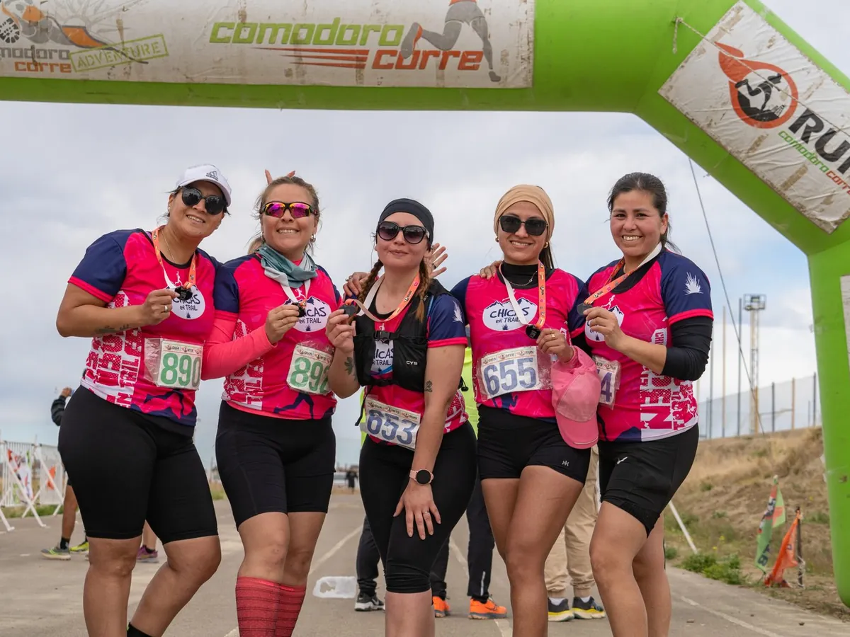 Cinco corredoras con remeras rosas full sublimadas y medallas bajo el arco de llegada de Comodoro Corre